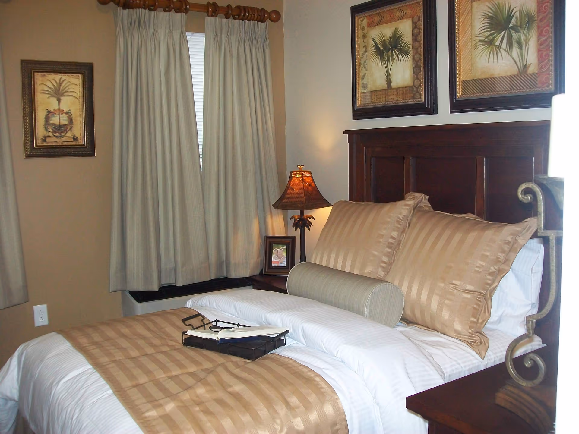 A neatly made bed with beige and white striped bedding, two large pillows, and a cylindrical bolster pillow in a bedroom. There is a wooden headboard, a nightstand with a decorative lamp, a framed photo, and two framed pictures of palm trees on the wall. Beige curtains cover the window.