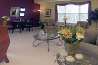 A cozy living room with beige carpet and walls painted in cream and purple. The room features a glass coffee table with a decorative glass pitcher and glasses, a side table with a flower arrangement, two armchairs near a large window with blinds, a floor lamp, and a desk with a chair and framed pictures on the wall.