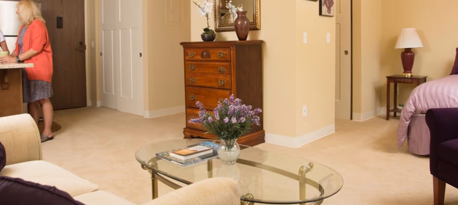 Interior view of a senior living residence showing a cozy living area with a glass coffee table holding a vase of purple flowers and some books. To the right, there is a bed with a purple bedspread and a bedside table with a lamp. A wooden chest of drawers with decorative items and a mirror is against the wall. A woman in a coral cardigan and patterned dress is standing near a kitchen counter on the left side of the image.