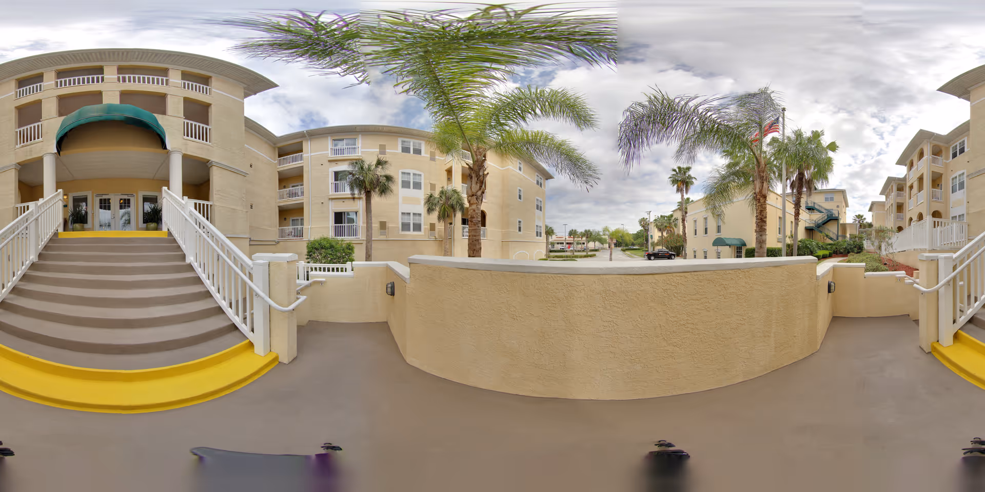 Outdoor view of a senior living facility showing a beige multi-story building with balconies, palm trees, and a staircase with white railings and yellow safety markings leading to an entrance with a green awning. The sky is partly cloudy.