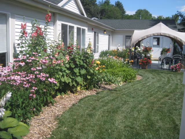 Well-maintained grassy courtyard alongside a single-story assisted living building with colorful flower beds and a covered patio seating area.