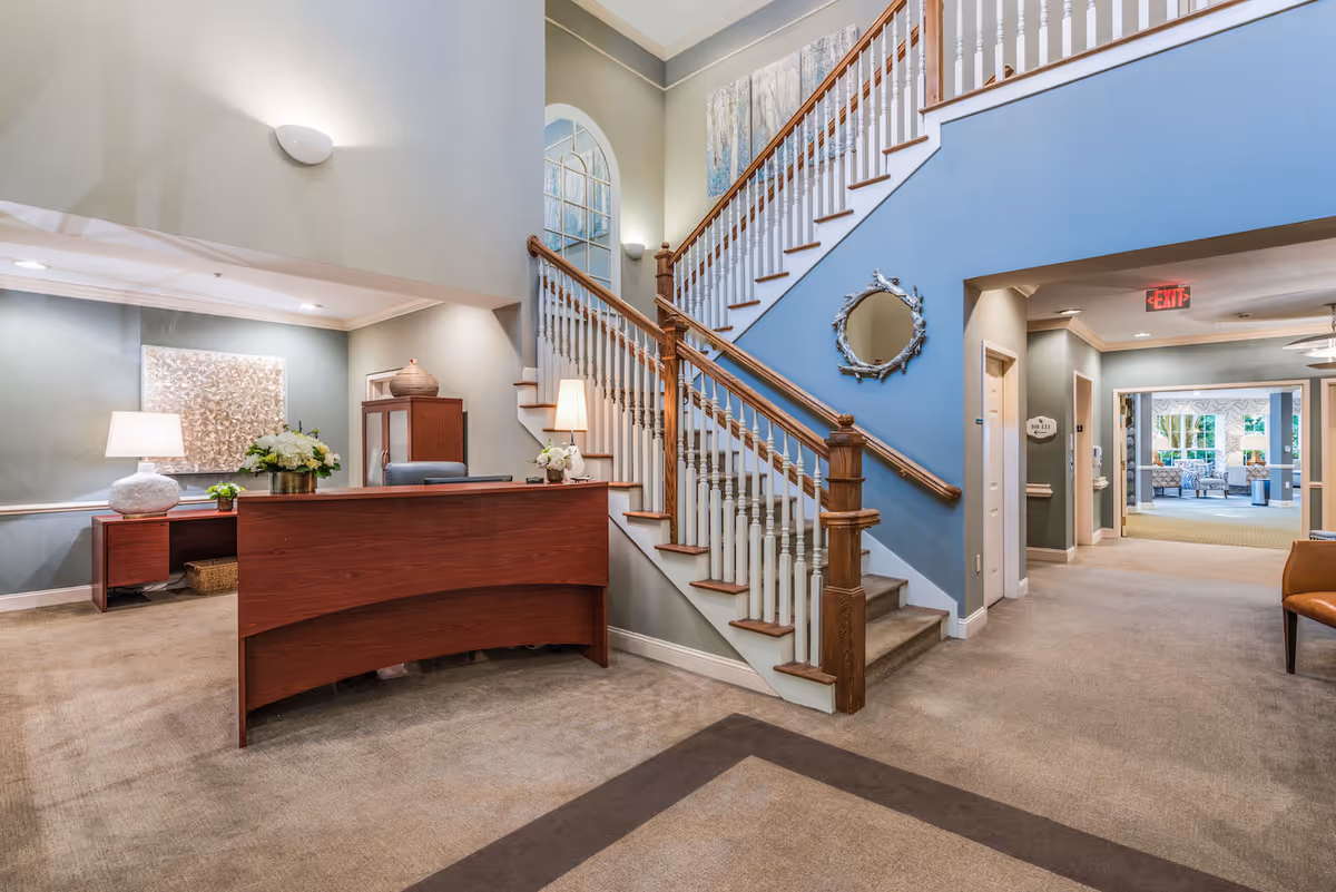 Interior view of a senior living facility lobby with a wooden reception desk, a staircase with wooden handrails and white balusters, a decorative mirror on a blue accent wall, and a hallway leading to a seating area with large windows.