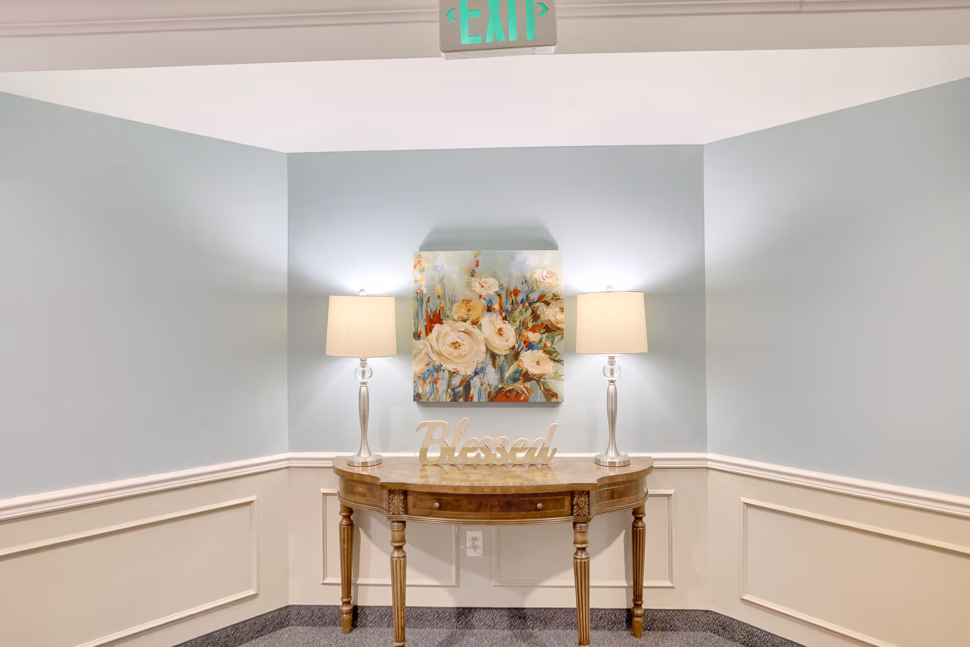 A small wooden console table with curved front and four legs is placed against a light blue wall with white wainscoting. On the table are two matching lamps with beige shades and a decorative sign that says 'Blessed'. Above the table hangs a colorful floral painting. An illuminated green exit sign is visible above the wall.