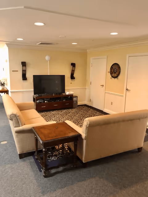 A cozy living room area with two beige sofas facing a flat-screen TV on a wooden TV stand. The room has a patterned area rug, a wooden side table between the sofas, and light yellow walls with white trim. There are two closed white doors on the right wall and decorative wall sconces and a circular wall decoration.