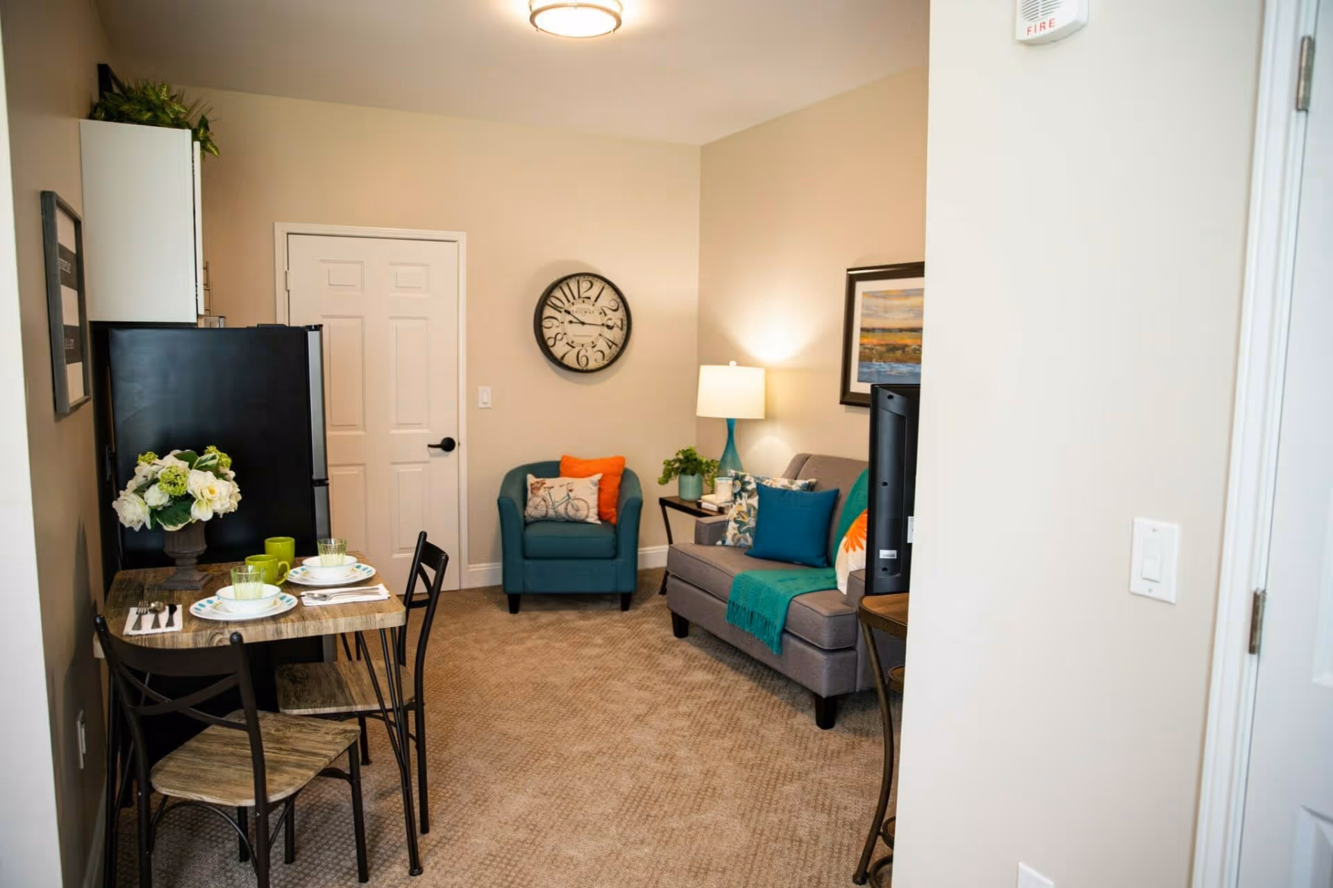 Small furnished living area with a sofa, armchair, lamp, wall clock, and a small dining table beside a refrigerator.