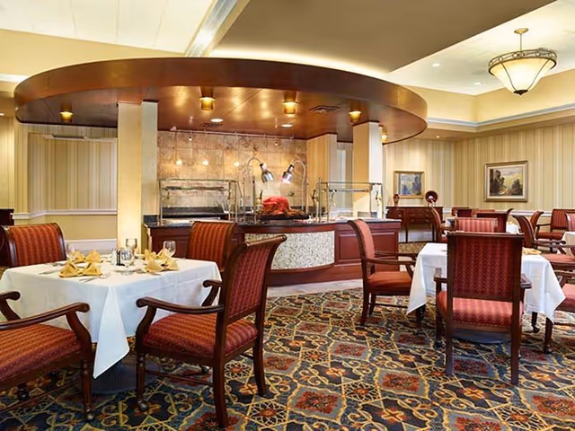 Elegant dining room with a central buffet station, tables set with white linens and upholstered chairs under warm lighting.