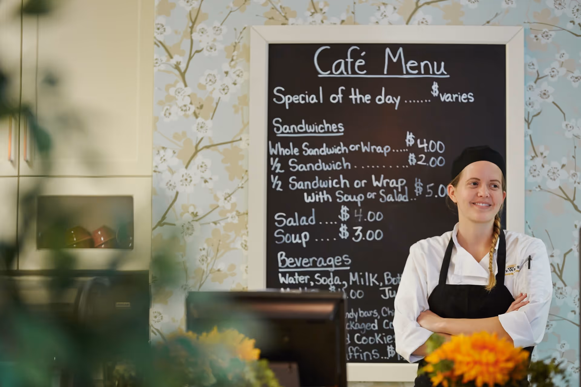 A female chef wearing a white uniform and black apron stands with arms crossed in front of a chalkboard menu in a cafe. The menu lists specials, sandwiches, salads, soups, and beverages. The background features floral wallpaper and kitchen cabinets.