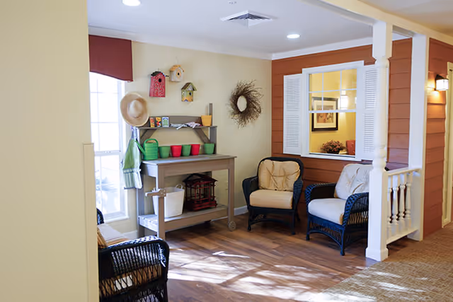 A cozy indoor seating area with two cushioned wicker chairs and a small table with gardening supplies including colorful pots, watering cans, and birdhouses. The space has warm lighting, wooden flooring, and a window with white shutters looking into another room.