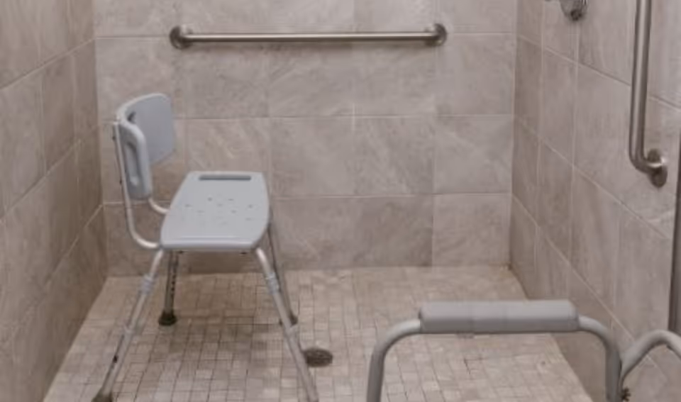 Accessible shower area with beige tiled walls and floor, equipped with a gray shower chair with backrest, grab bars on the walls, and a handheld showerhead.
