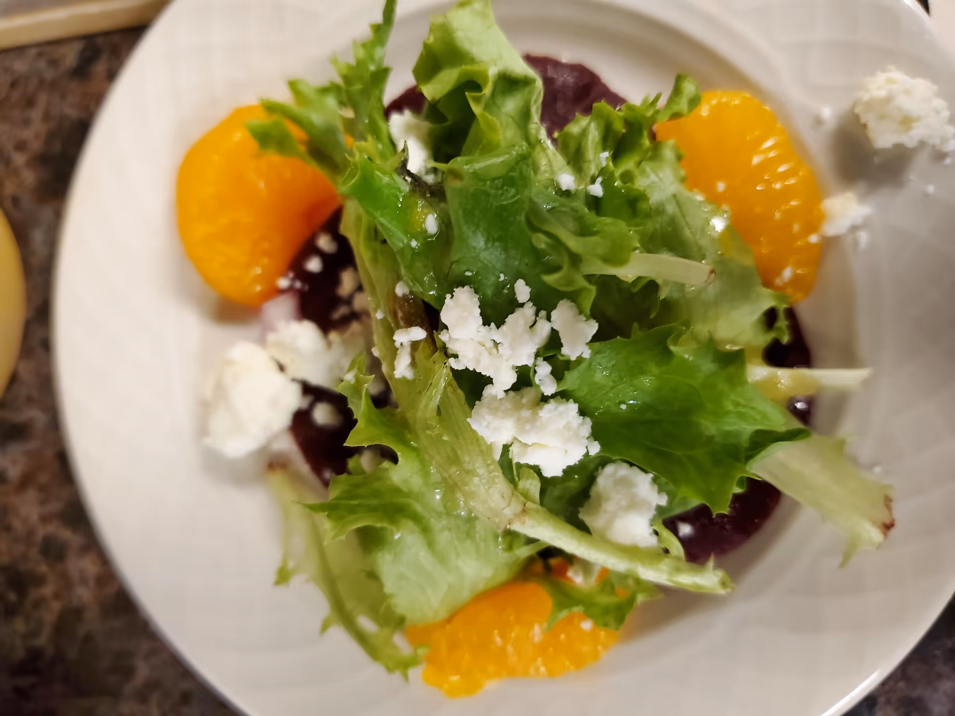 A close-up view of a salad on a white plate, featuring leafy greens, crumbled white cheese, slices of mandarin oranges, and beetroot slices underneath.