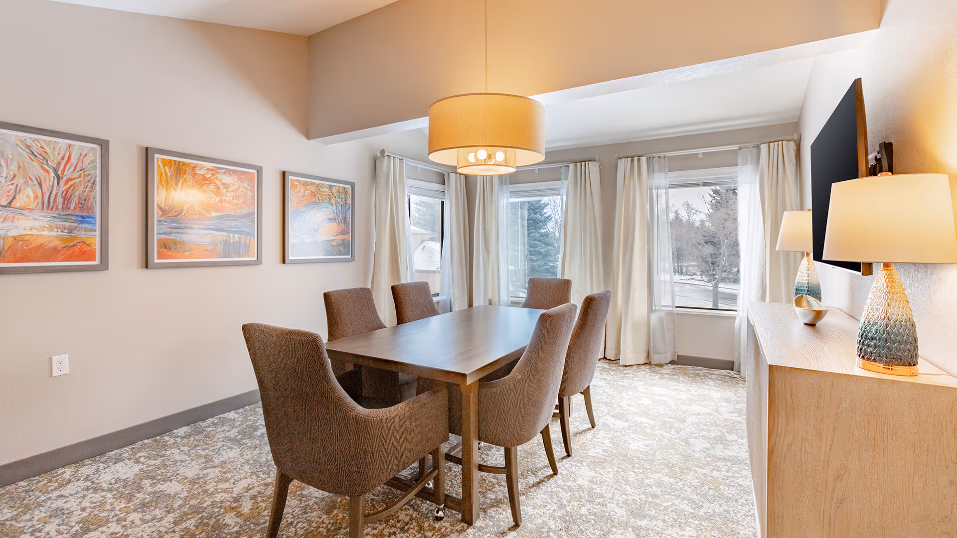 A bright dining room with a rectangular wooden table surrounded by six upholstered chairs. The room features large windows with sheer curtains allowing natural light to fill the space. On one wall, there are three colorful landscape paintings, and on the opposite side, a wooden sideboard with two decorative lamps and a mounted flat-screen TV above it. A modern ceiling light fixture hangs above the table.
