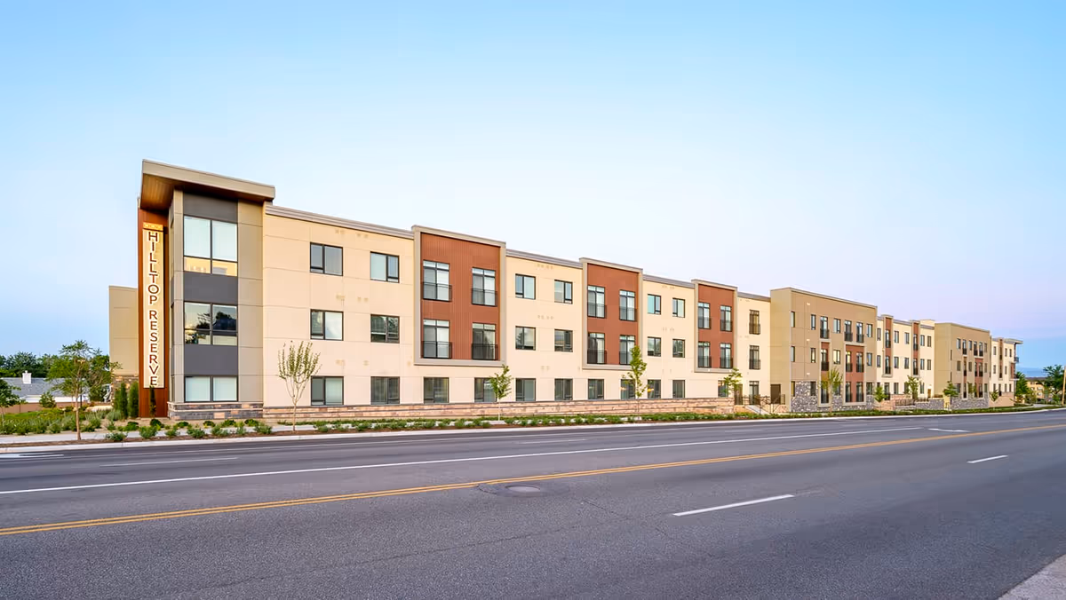 Exterior view of a modern three-story senior living facility named Hilltop Reserve Senior Living, with a clean facade featuring beige and brown panels, multiple windows, small trees, and landscaping along the sidewalk beside a wide road.