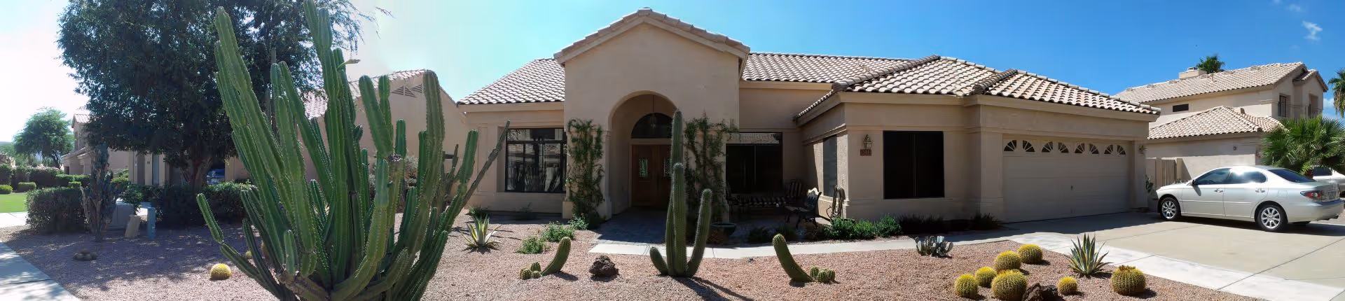 Front exterior view of a single-story assisted living facility building with a tiled roof, desert landscaping featuring various cacti and shrubs, a driveway with a parked silver car, and a clear blue sky.