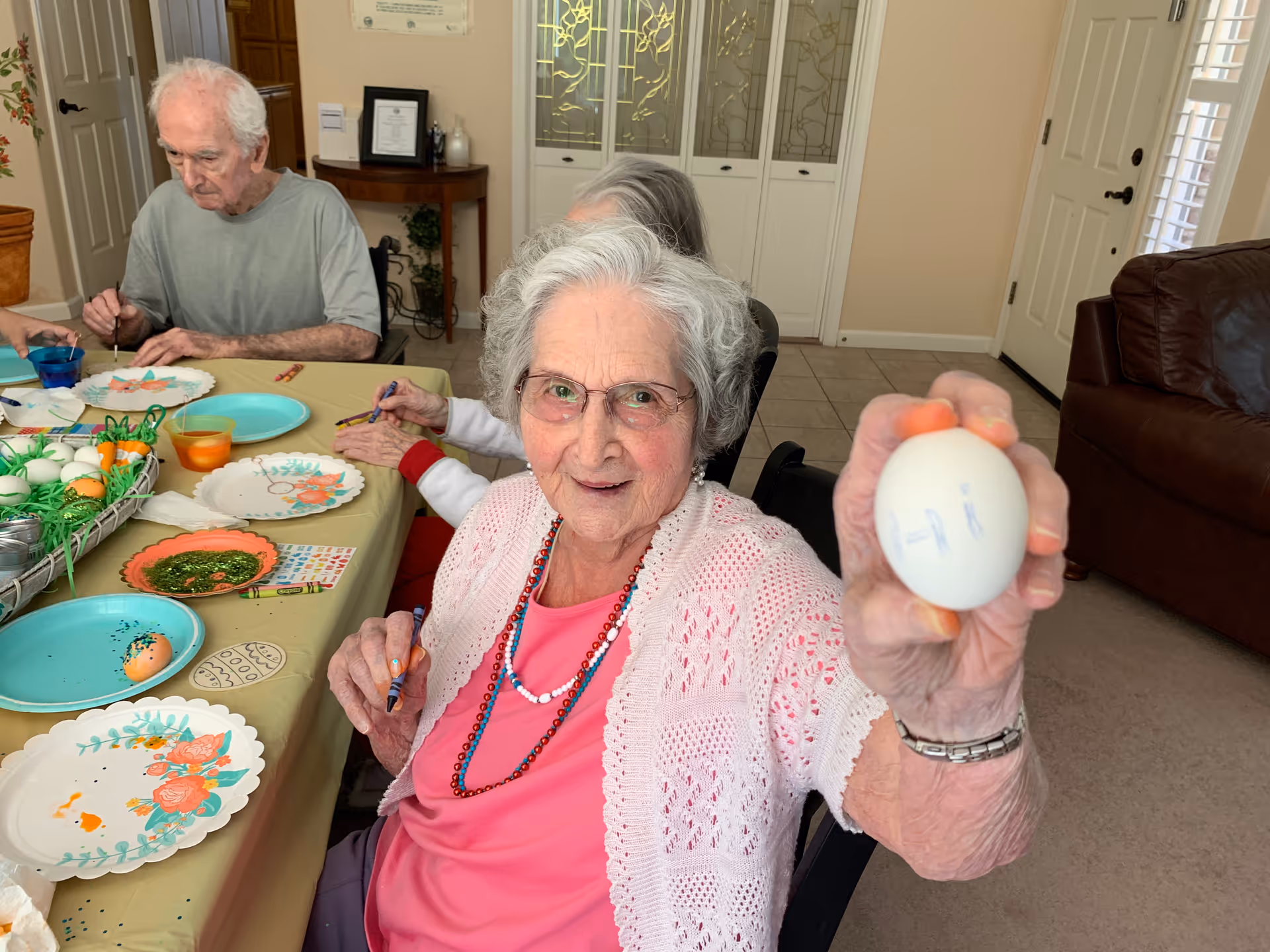 An elderly woman with gray hair and glasses is sitting at a table in a living room, holding up a decorated egg with the word 'Hi' written on it. She is smiling and wearing a pink shirt with a white knitted cardigan and beaded necklaces. Other elderly individuals are seated around the table, engaged in decorating eggs with various art supplies and plates on the table. The room has beige walls, a brown leather couch, and a door with glass panels in the background.
