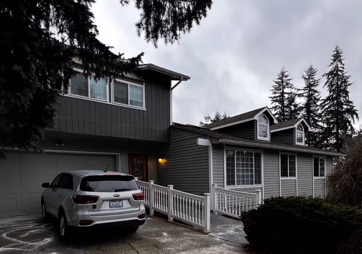 Exterior view of a two-story residential building with gray siding and white trim. There is a white car parked in the driveway in front of a garage. The building has multiple windows, including two dormer windows on the upper floor. Trees and bushes surround the property under a cloudy sky.
