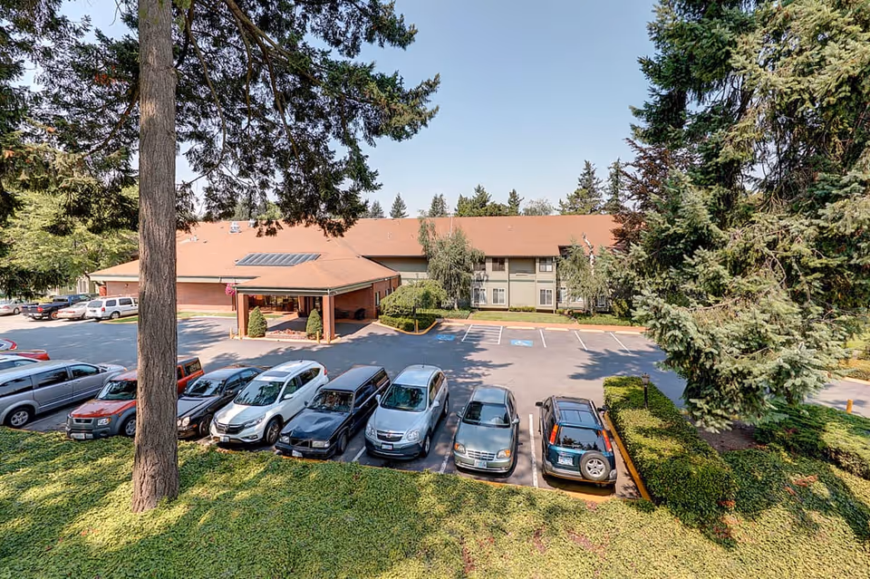 Exterior view of Firwood Gardens facility showing a parking lot with several parked cars in front of a two-story building with a red roof, surrounded by tall trees and greenery under a clear blue sky.