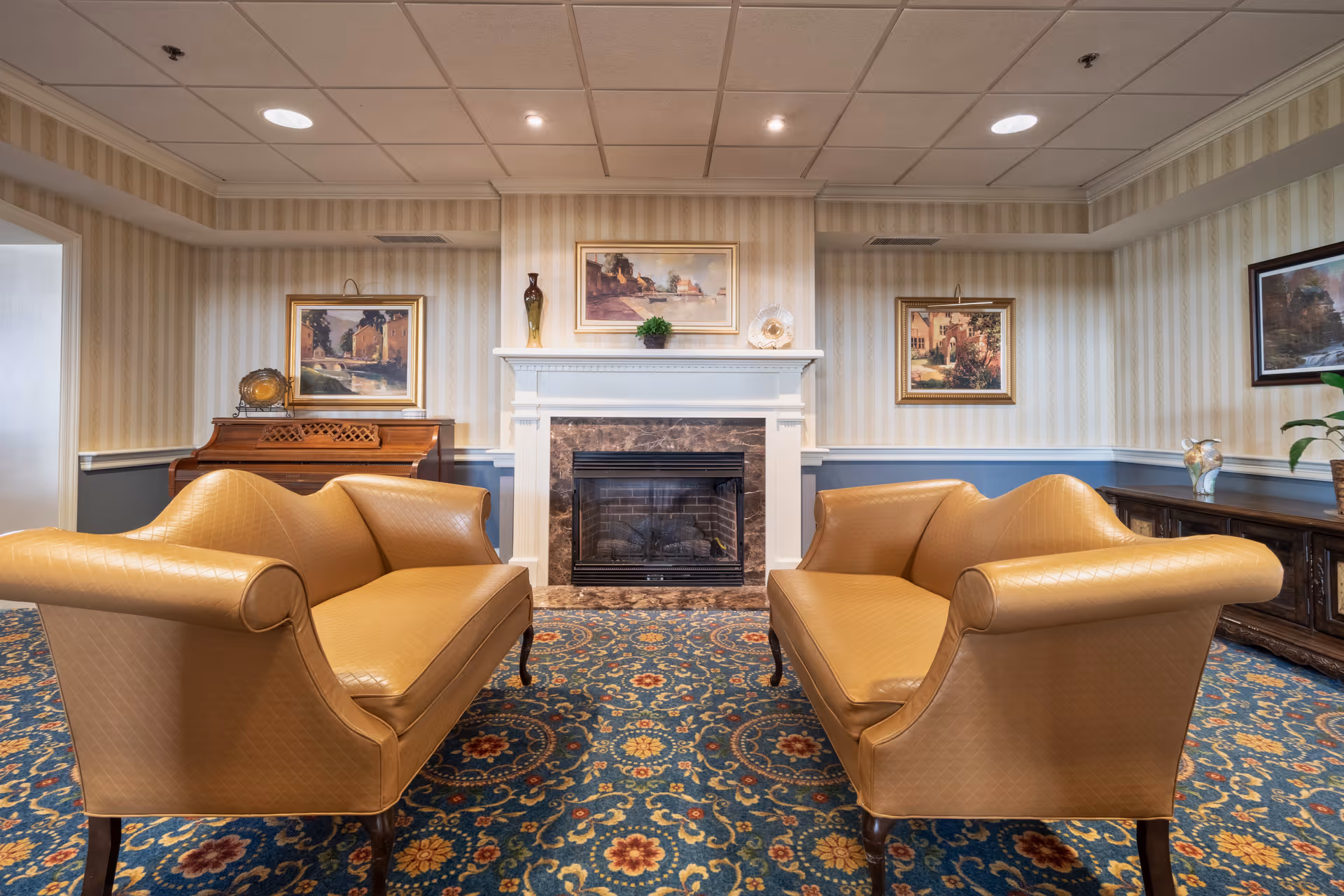 A cozy living room with two tan leather sofas facing each other on a patterned carpet. In the center background is a white fireplace with a dark marble surround, decorated with a small plant and a decorative item. The walls have striped wallpaper with framed paintings hanging above a piano on the left and a wooden cabinet on the right.