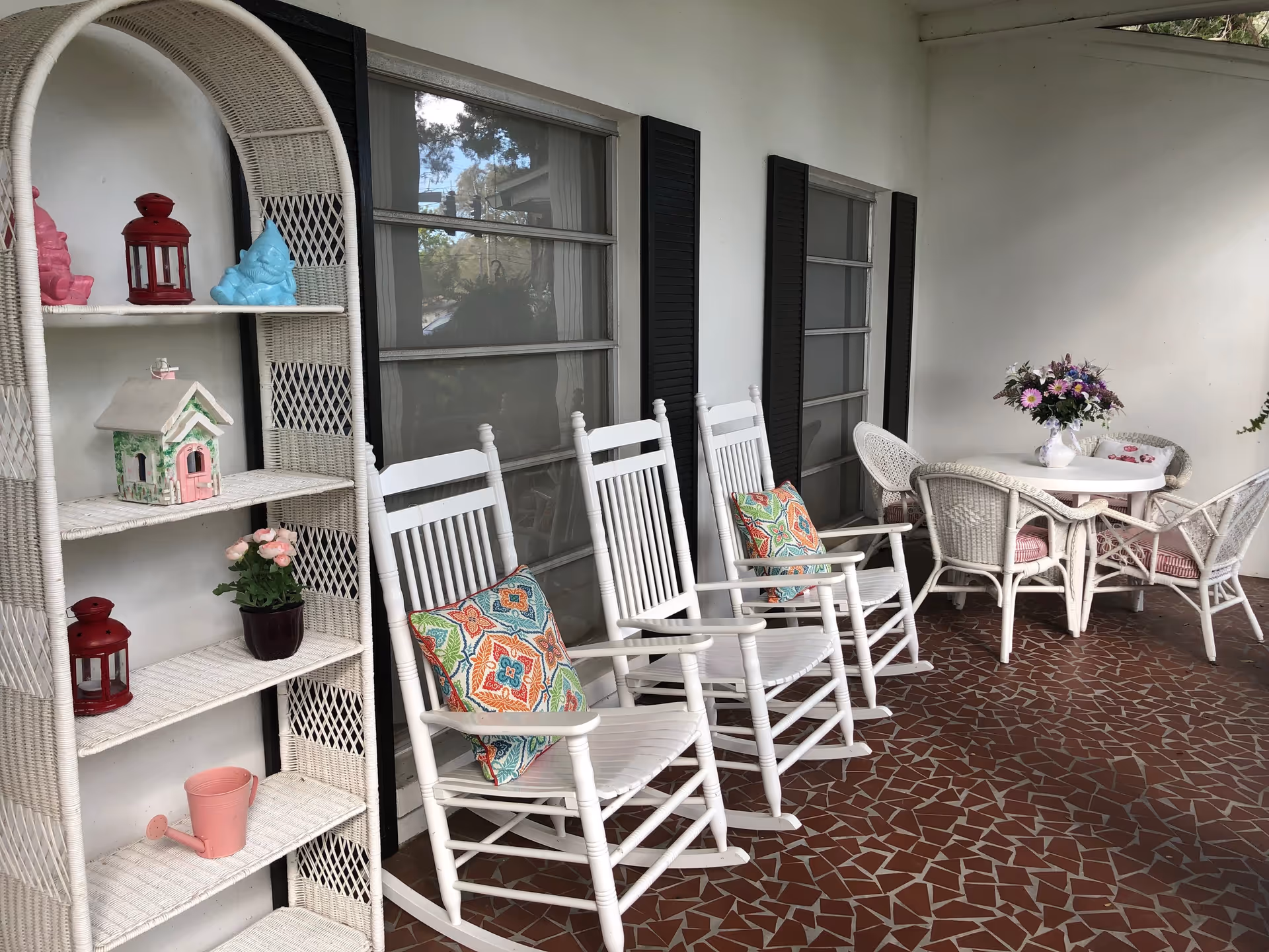 A covered outdoor patio area with three white rocking chairs each with a colorful patterned cushion, a white wicker shelf holding decorative items including a small birdhouse, lanterns, and plants, and a white round table with four wicker chairs and a vase of flowers on top. The floor is tiled with a red and white mosaic pattern.