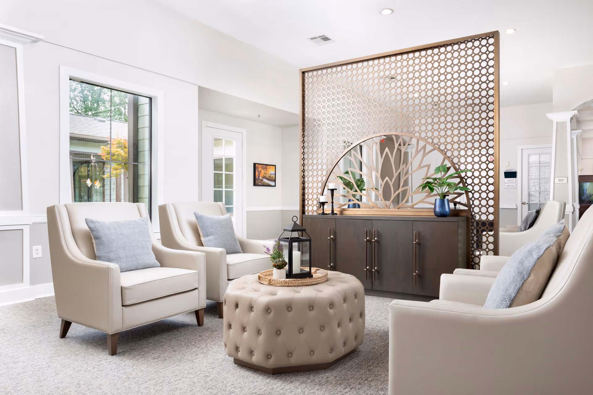 A bright and modern living room area with four beige armchairs arranged around a tufted ottoman. The ottoman has a tray with a lantern and small plants. Behind the seating area is a decorative wooden screen with a circular mirror featuring a tree design. The room has large windows and light-colored walls.