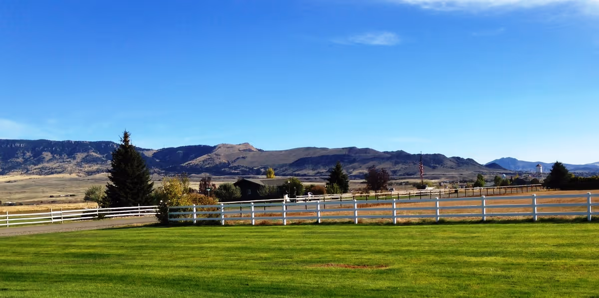 A wide open outdoor area with a well-maintained green lawn, white wooden fences, scattered trees, and a backdrop of mountains under a clear blue sky.