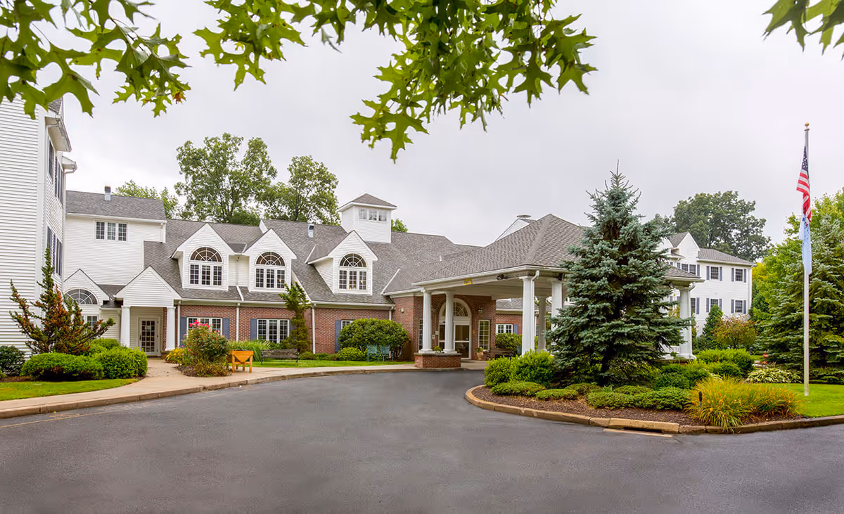 Exterior view of Middlebrook Farms at Trumbull, showing a large building with white siding and brick accents, multiple windows with arched tops, a covered entrance with white columns, landscaped greenery including bushes and trees, and an American flag on a flagpole to the right.
