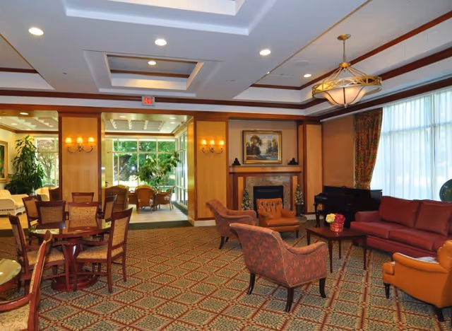 A spacious and well-lit senior living facility common area featuring a patterned carpet, multiple seating arrangements including upholstered armchairs and a sofa, a wooden table with chairs, a fireplace with a painting above it, large windows with curtains allowing natural light, and a piano in the corner. The room has warm wood paneling and ceiling lights.