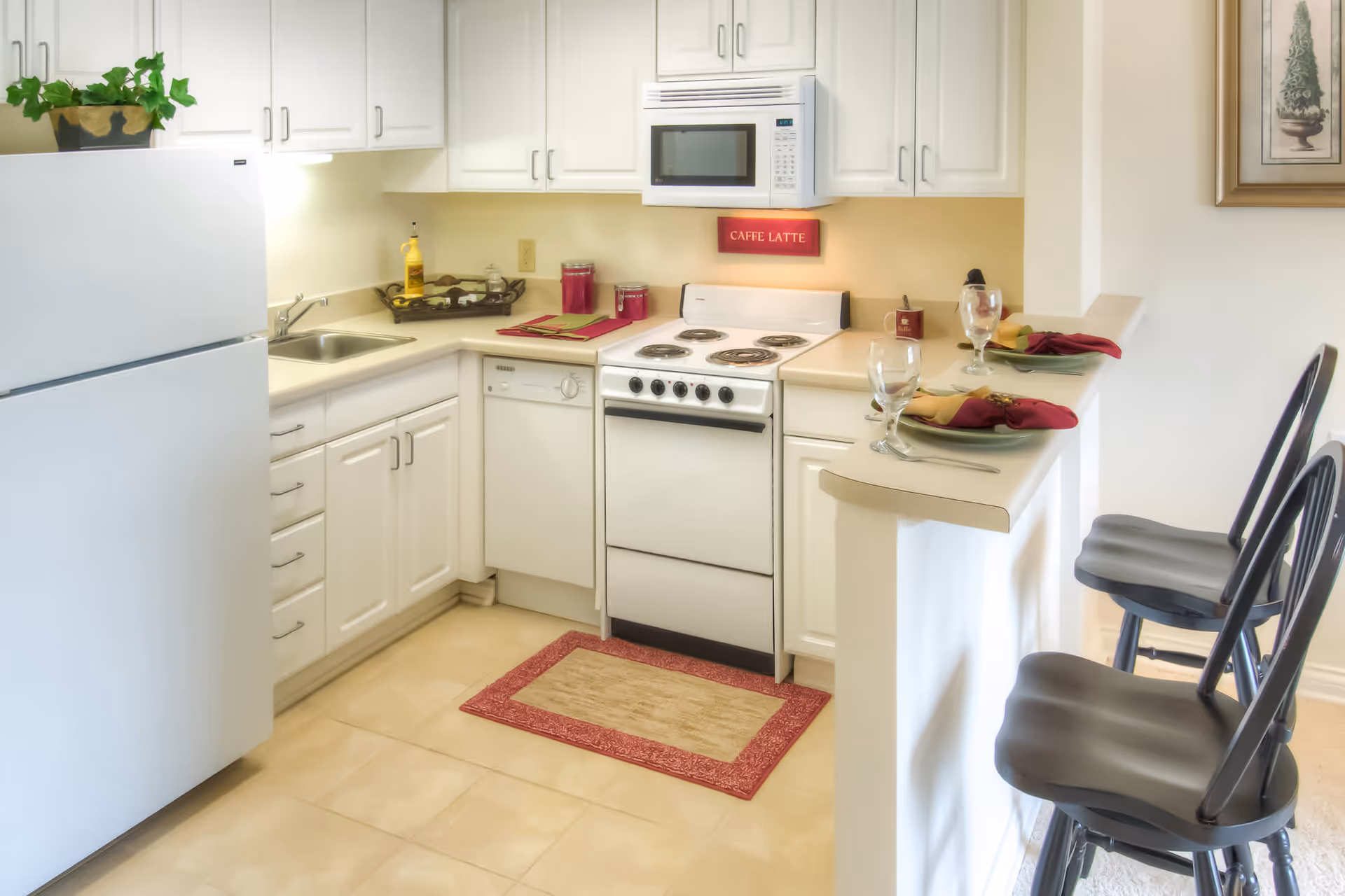 A clean, bright kitchen with white cabinets and appliances including a refrigerator, stove, microwave, and dishwasher. There is a small red and beige rug in front of the stove, a plant on top of the refrigerator, and a counter with two place settings with plates, napkins, and glasses. A red sign above the stove reads 'CAFFE LATTE'.
