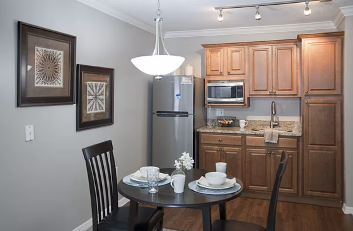 Small kitchen and dining area with wooden cabinets, a stainless refrigerator, a round table set for two, and a hanging light.
