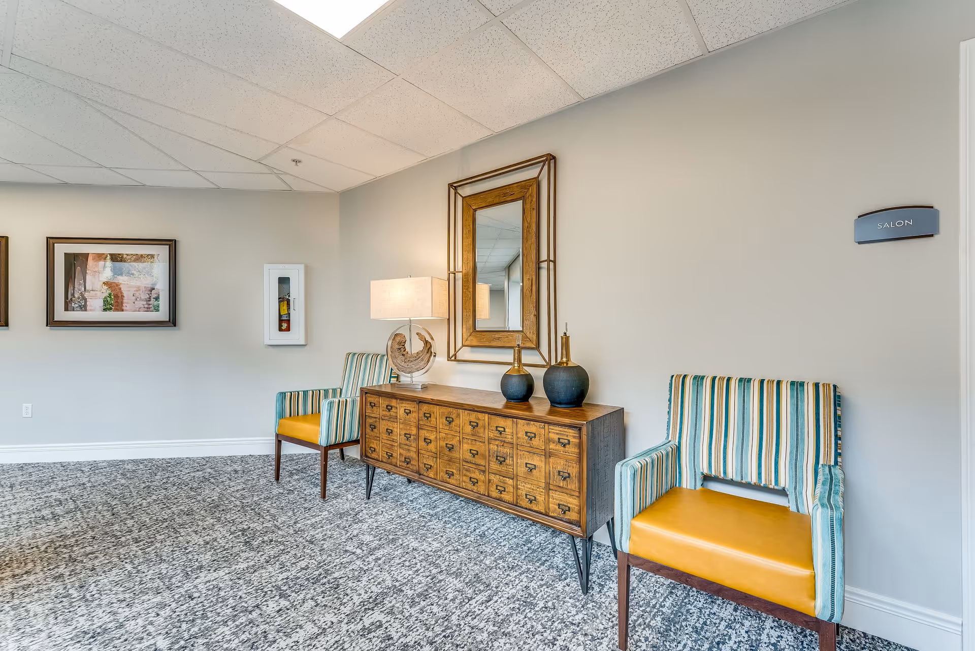 A waiting area with two striped armchairs with yellow seats, a wooden sideboard with multiple drawers, a table lamp, decorative vases, and a large framed mirror above it. The wall has a sign labeled 'SALON' and two framed pictures are visible on the adjacent wall. The floor is carpeted and the ceiling has white tiles.