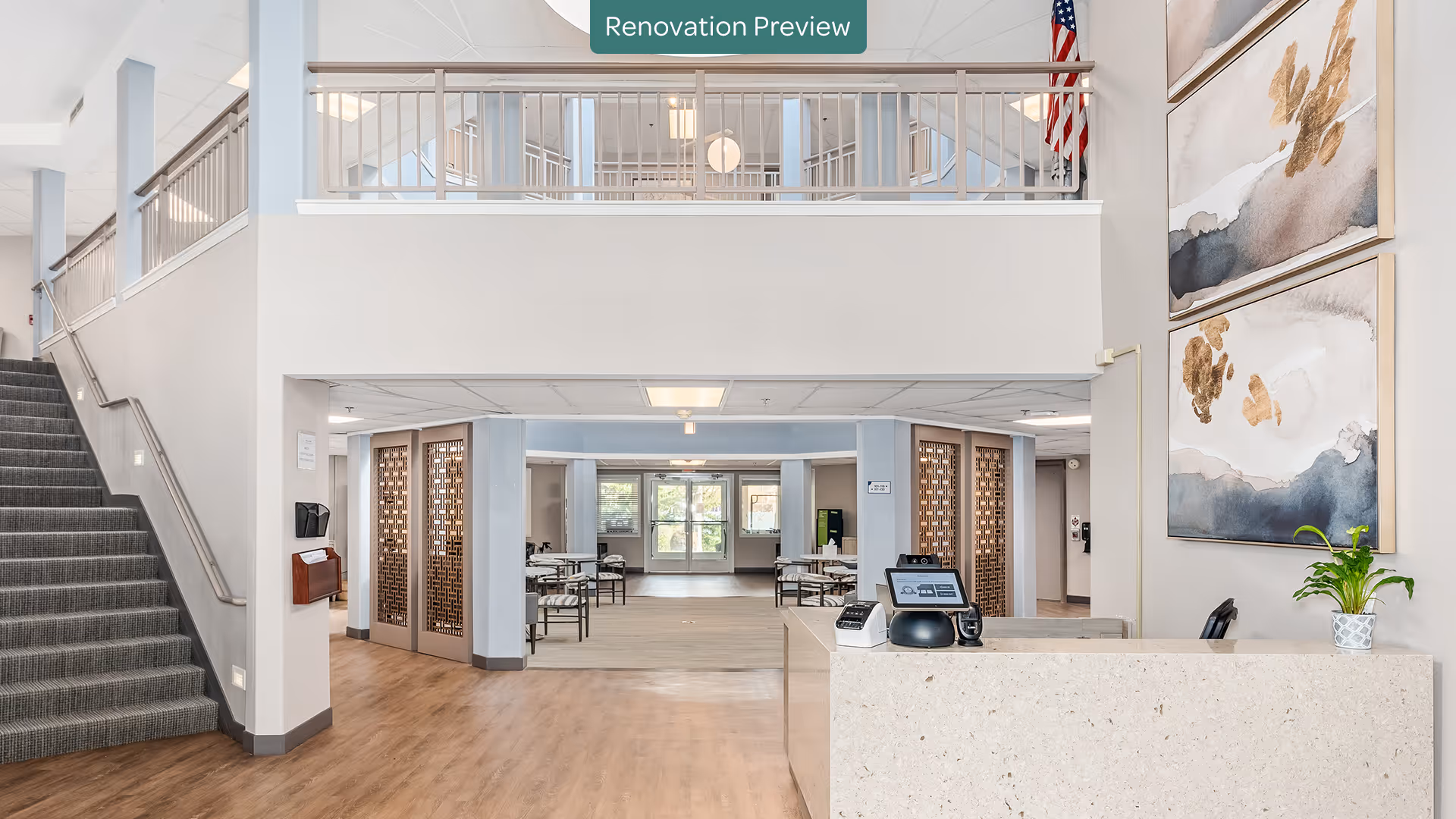 Interior view of a senior living facility lobby area with a reception desk on the right, a staircase on the left, and a seating area with tables and chairs in the background. The walls are decorated with abstract paintings and an American flag is visible near the ceiling. A sign above reads 'Renovation Preview'.