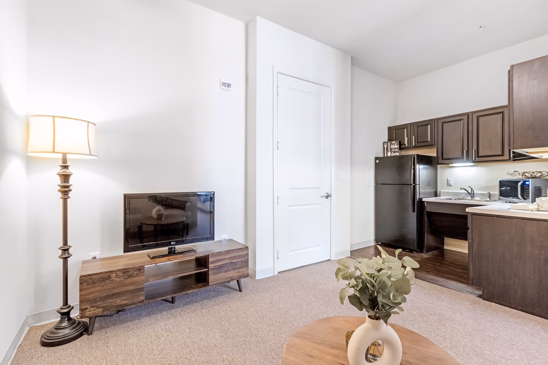 Interior view of a living space featuring a wooden TV stand with a flat-screen TV, a tall floor lamp with a beige shade, a round wooden coffee table with a white vase and green foliage, and a kitchen area with dark wood cabinets, a black refrigerator, a microwave, and a sink.