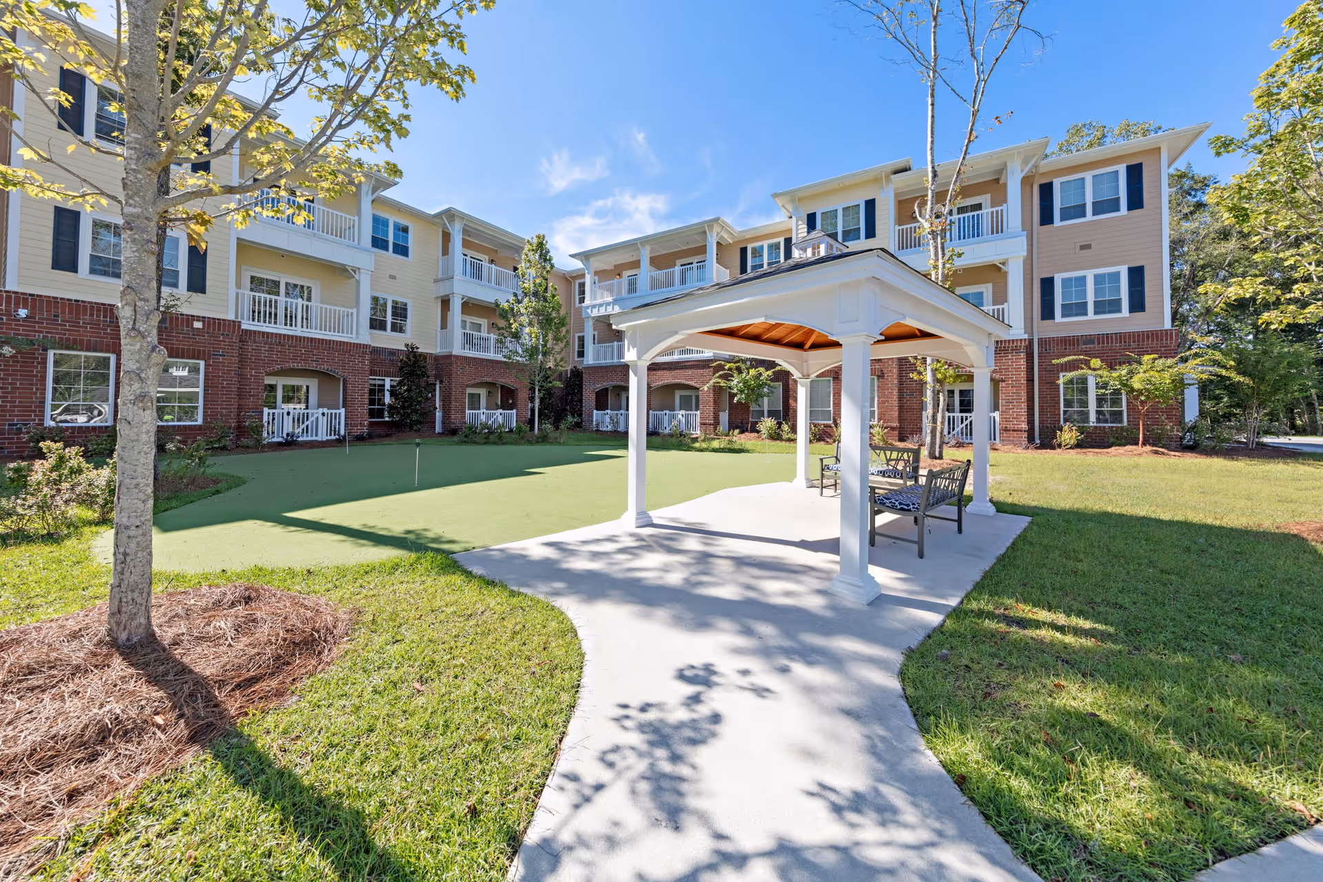 Outdoor view of a senior living facility with a three-story building featuring balconies and large windows. In the foreground, there is a white gazebo with benches underneath, surrounded by a well-maintained lawn and trees. The sky is clear and blue.