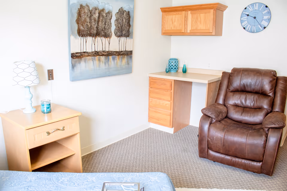 A cozy corner of a senior living room featuring a brown leather recliner, a small wooden desk with drawers and a countertop, a wooden nightstand with a lamp and a candle, a wall clock, and a painting of trees by a lake on the wall.