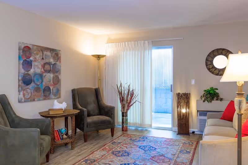 A cozy living room with two gray armchairs and a beige sofa with red cushions. There is a wooden side table with books and a decorative white bird, a floor lamp, a colorful patterned rug, a tall vase with dried plants, a round mirror on the wall, and a sliding glass door with sheer curtains leading outside.