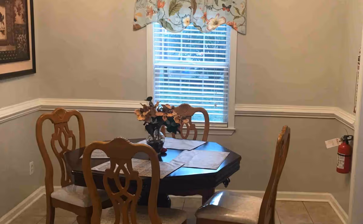 A small dining area with a wooden table and four matching chairs. The table has placemats and a vase with artificial flowers. There is a window with white blinds and a floral valance above it. A fire extinguisher is mounted on the wall to the right, and a framed picture hangs on the left wall.