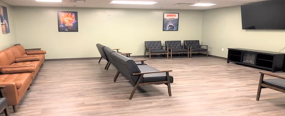Large common lounge with rows of mid-century style chairs and sofas facing a wall-mounted TV, posters on pale green walls, and wood-look flooring.