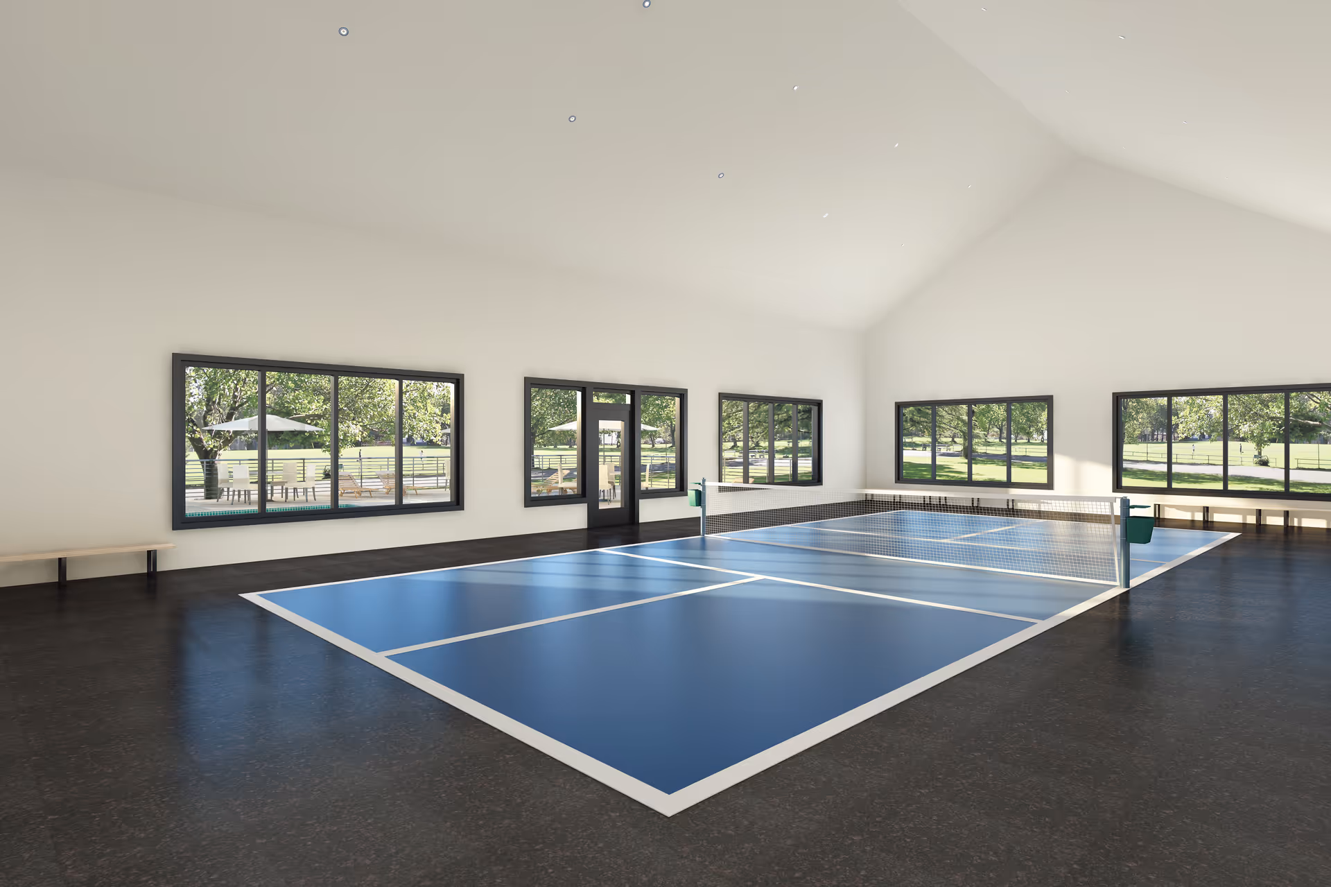 Indoor room with a blue pickleball court marked on the floor, surrounded by large windows showing a green outdoor area with trees and benches. The room has a high ceiling and benches along the walls.