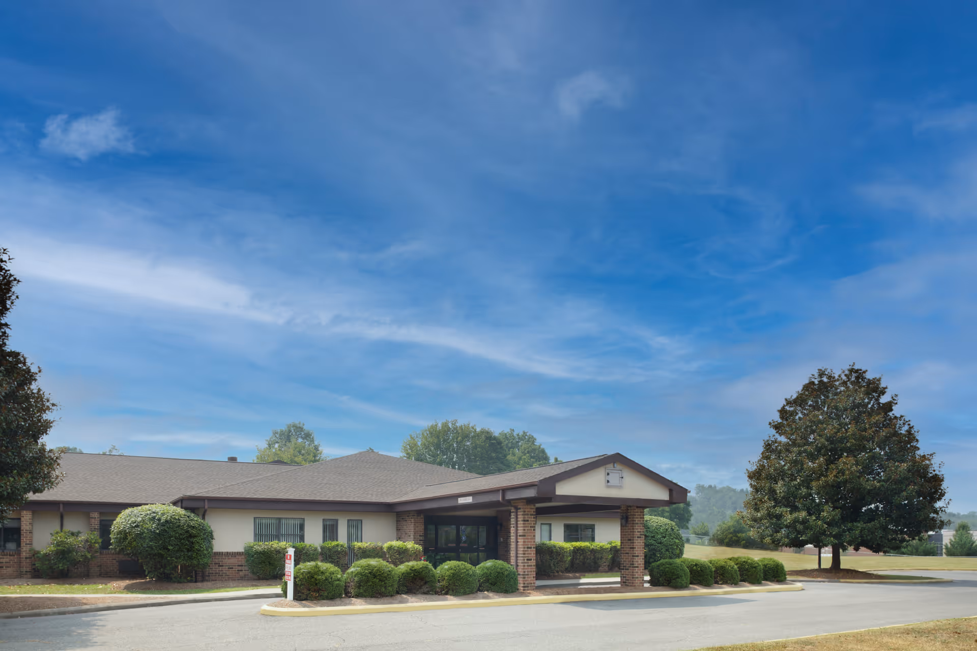 Single-story senior living building with a covered entrance, trimmed shrubs, and a large tree under a blue sky.