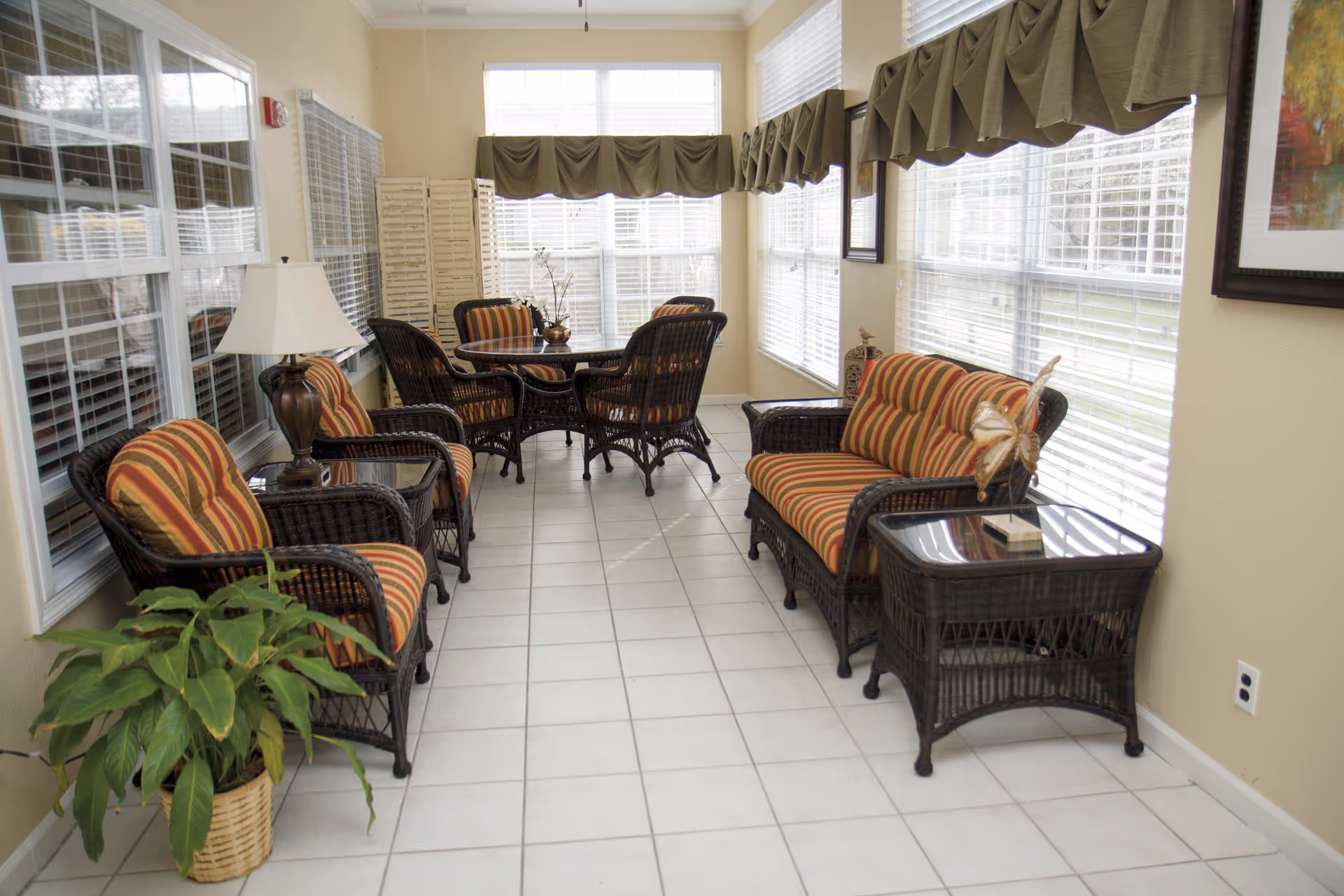 A bright sunroom with large windows on two sides, furnished with dark wicker chairs and a loveseat with striped orange and yellow cushions. There is a round table with four chairs in the background, a side table with a butterfly decoration, a lamp on a small table, and a potted plant on the tiled floor.