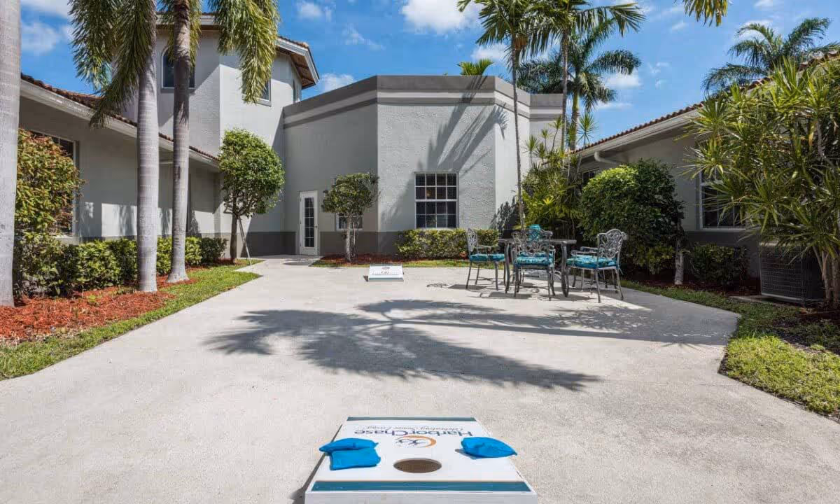 Outdoor patio area at HarborChase of Tamarac with a concrete surface, a cornhole game set up, metal table and chairs with blue cushions, surrounded by palm trees and bushes under a clear blue sky.