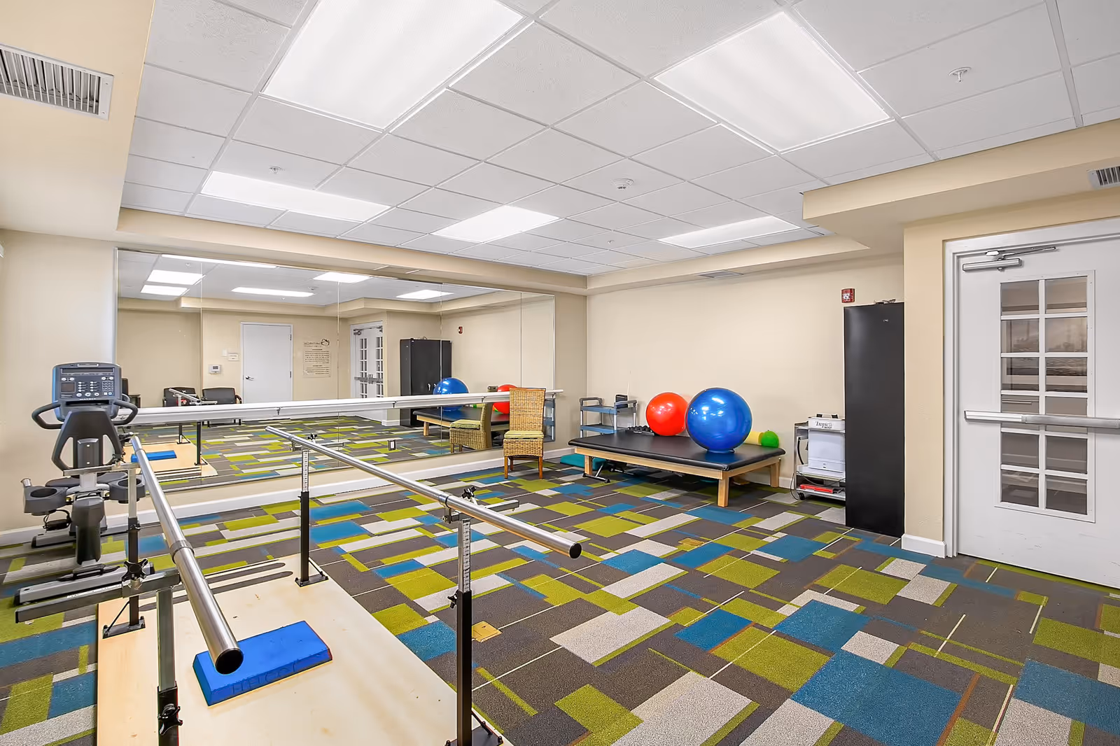 A bright exercise room with colorful patterned carpet, a large wall mirror, parallel bars for walking support, an exercise bike, a bench with large red and blue exercise balls, and a wicker chair. The room has a white ceiling with fluorescent lights and a white door with glass panels.