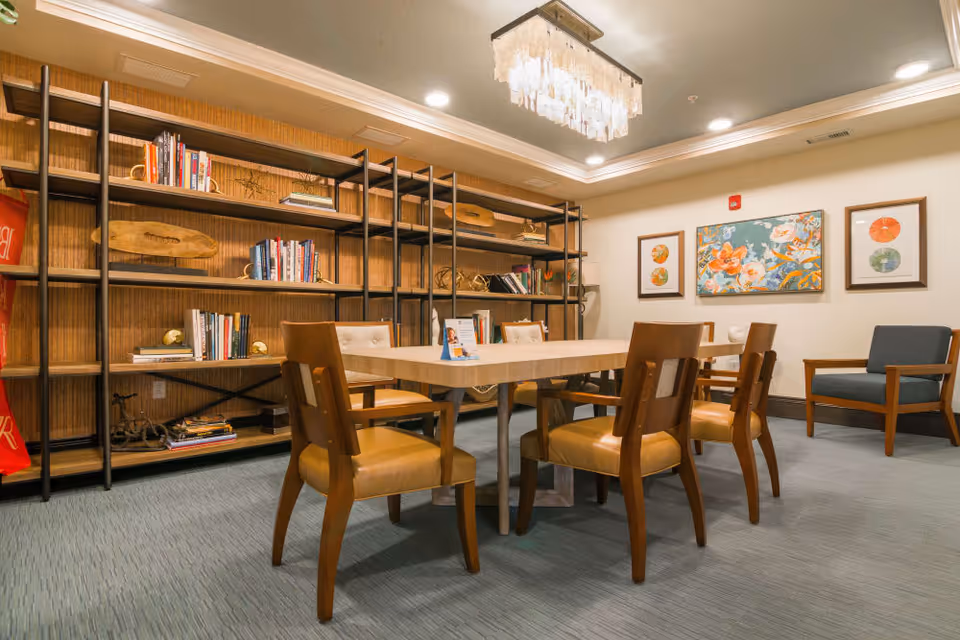 Well-lit communal room with a central table surrounded by wooden chairs, bookshelves along the wall, and framed artwork on the wall.