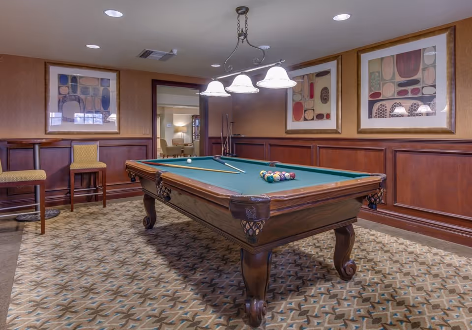 Interior room with a pool table in the center, two pool cues resting on the table, and a rack of billiard balls arranged for a game. The room has patterned carpet, wood-paneled walls with three framed abstract artworks, two yellow cushioned chairs, and a hanging light fixture above the pool table. An open doorway leads to another room with seating visible.