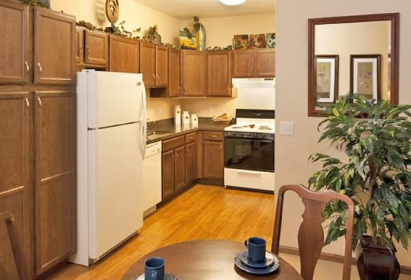 A kitchen area with wooden cabinets, a white refrigerator, dishwasher, and stove. The floor is wooden, and there is a small round dining table with two place settings in the foreground. A large green plant and a mirror are visible on the right side of the image.