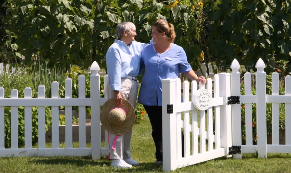 An elderly woman holding a straw hat with a pink ribbon stands next to a caregiver in a blue uniform. They are smiling at each other while standing by a white picket fence gate in a garden with tall green plants and sunflowers in the background.