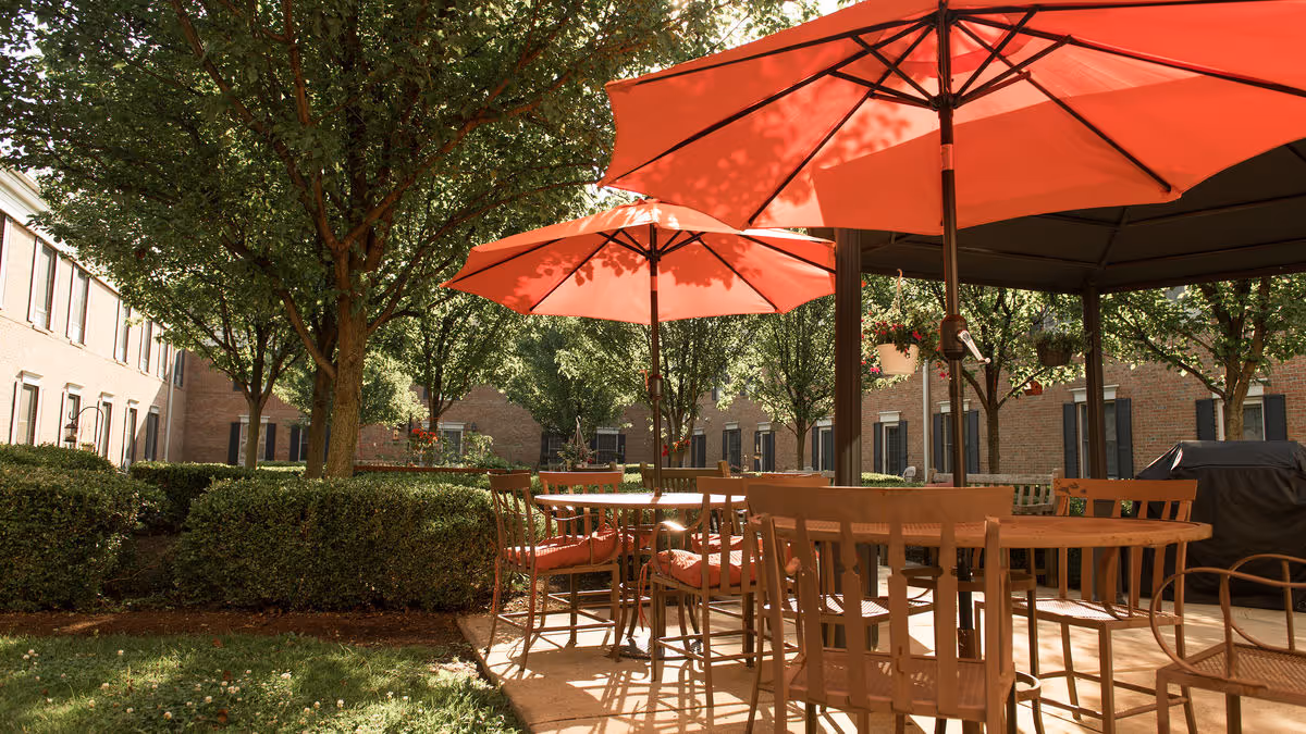 Sunlit courtyard with outdoor tables, chairs, and red umbrellas between brick residence buildings.