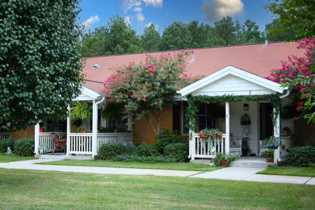 Single-story residential building with a red roof and white porch railings, surrounded by green trees and flowering bushes, with a concrete walkway and well-maintained lawn in front.