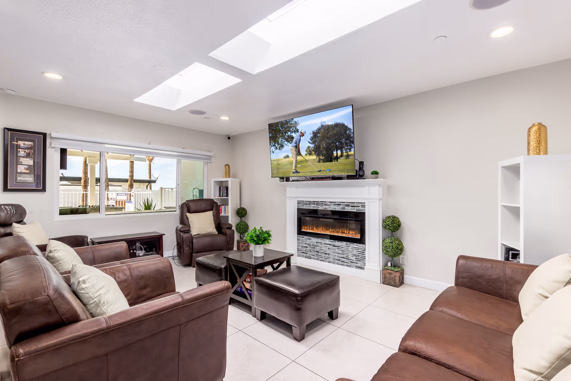 A bright living room with brown leather sofas and armchairs arranged around a dark wooden coffee table and ottoman. A modern electric fireplace with a tiled surround is mounted on the wall beneath a flat-screen TV showing a person playing golf. The room has white tiled floors, a large window with a view of palm trees outside, and two skylights in the ceiling. Decorative plants and shelves with books and ornaments complete the cozy setting.