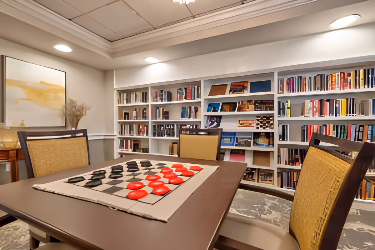 A cozy room with a table set for a game of checkers, surrounded by four chairs. Behind the table is a large built-in bookshelf filled with books and decorative items. The room has soft lighting from ceiling fixtures and a framed abstract painting on the wall.