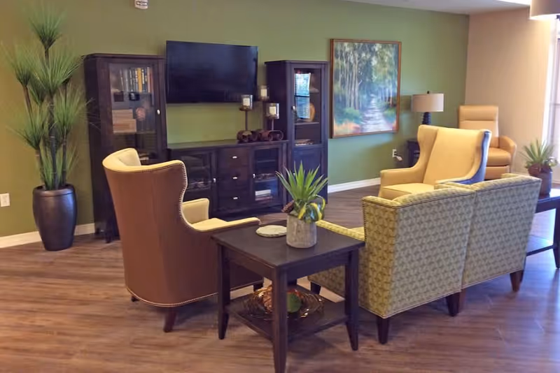 A cozy senior living room with green walls, wooden flooring, and comfortable seating including a patterned loveseat, two yellow armchairs, and a brown armchair. There is a dark wooden entertainment center with a flat-screen TV mounted above it, flanked by two glass-front cabinets. A side table with a potted plant is in the foreground, and a large painting of a forest path hangs on the wall. Additional plants and a floor lamp add to the warm ambiance.